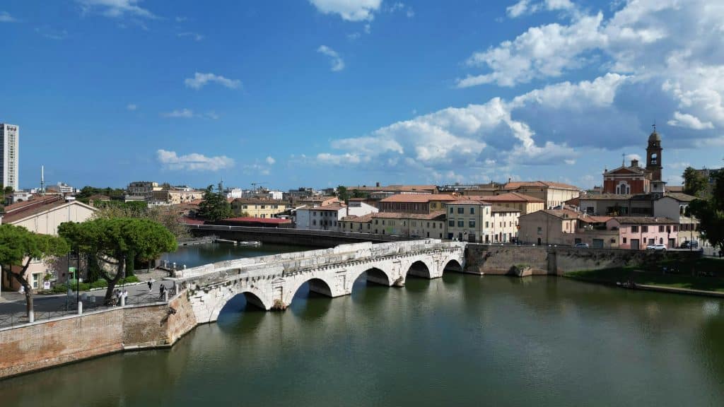 Puente de Tiberio en Rímini cruzando el río Marecchia, con edificios históricos al fondo y cielo azul.